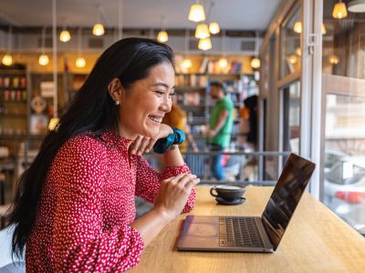 A modern young woman of Asian ethnicity, having a video call via laptop, at the modern and cozy cafeteria