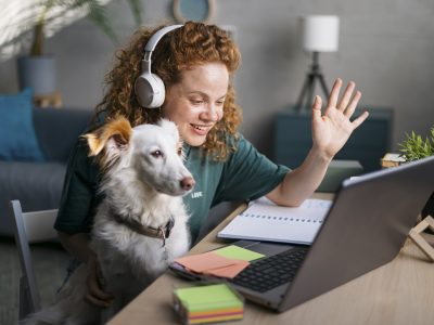 Happy young female freelancer casually working from the comfort of her home, attending a conference call using a laptop and waving to the screen while having her pet dog in her lap to keep her company